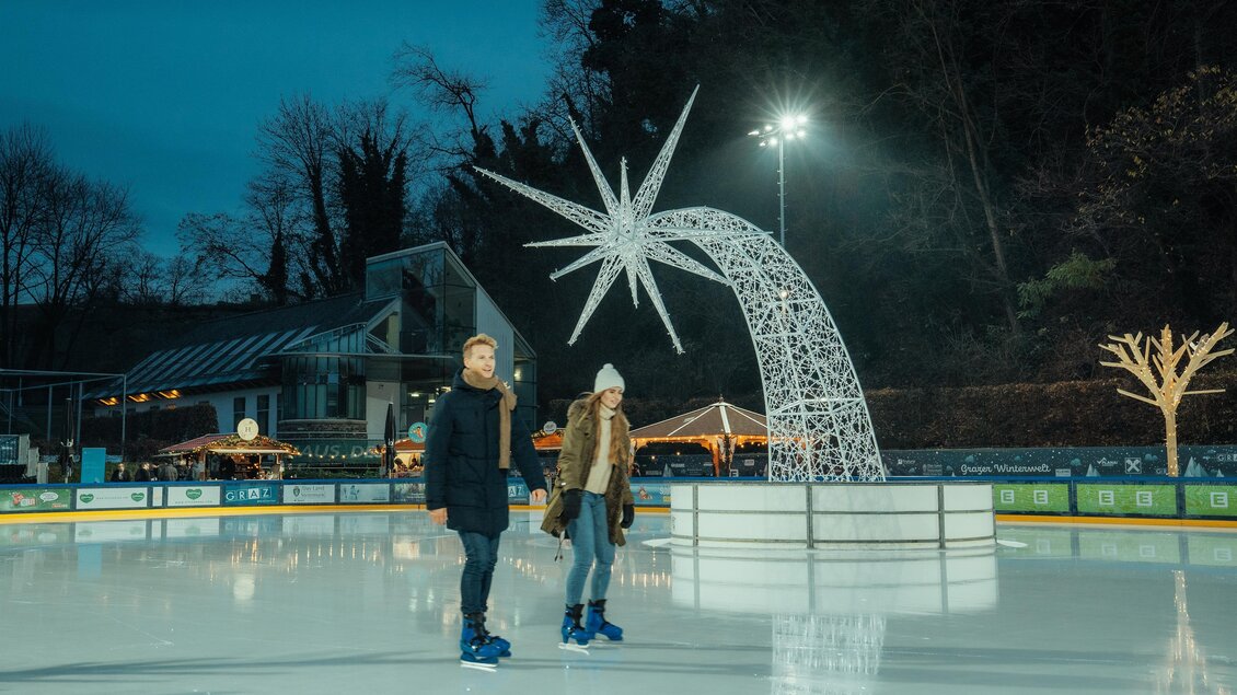 In der Abenddämmerung Eislaufen in der Grazer Winterwelt, bei Scheinwerferlicht. Der leuchtende Stern in der Winterwelt als Wahrzeichen.  | © Region Graz - studio draussen