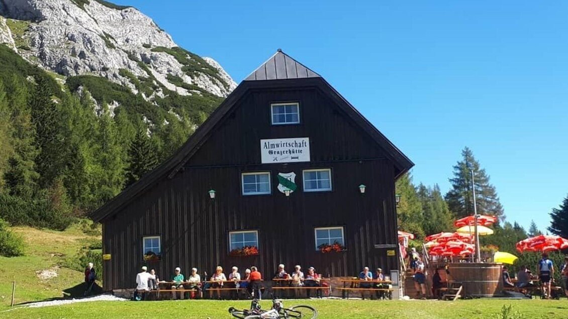 Eine gemütliche Almhütte umgeben von Bergen und Bäumen. Auf der Terrasse sitzen viele Gäste, während es sonnig und klar ist. | © Grazerhütte