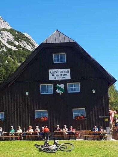 Eine gemütliche Almhütte umgeben von Bergen und Bäumen. Auf der Terrasse sitzen viele Gäste, während es sonnig und klar ist. | © Grazerhütte
