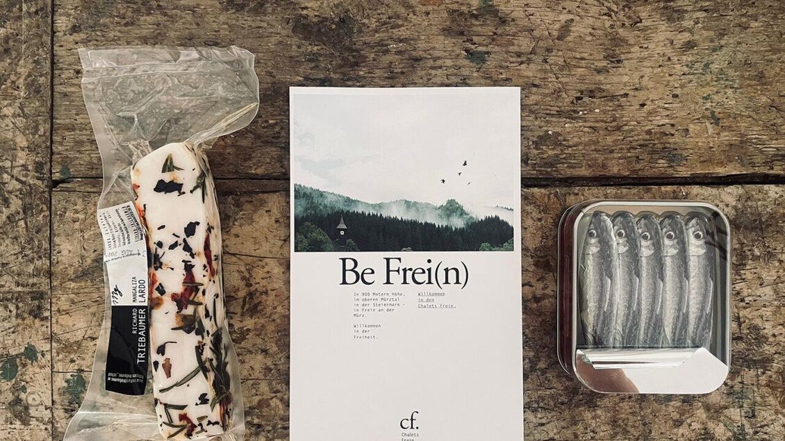 Eine Packung mit Snacks ist neben einem Heft mit dem Titel "Be Frei(n)" und einer Dose mit kleinen Fischen angeordnet. Der Hintergrund ist eine rustikale Holzoberfläche. | © Frein eleven ges.m.b.h.