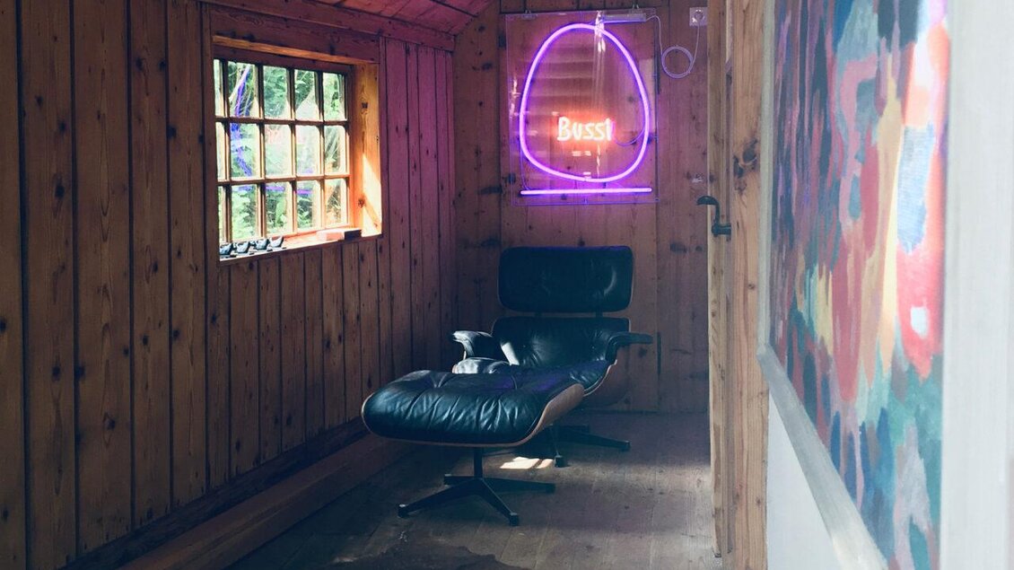 Ein gemütlicher Raum mit Holzverkleidung und einem modernen Sessel. Ein neonfarbener Schriftzug mit dem Wort "BASS" beleuchtet die Wand. | © Frein eleven ges.m.b.h.