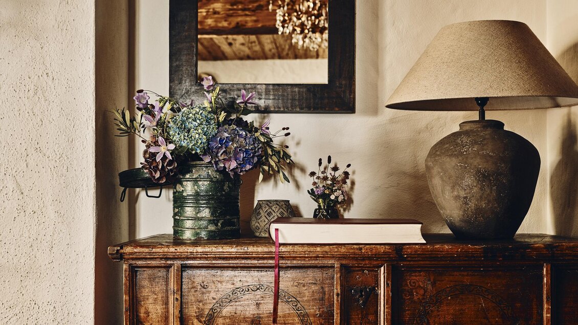 Ein rustikales Sideboard aus Holz mit einer Lampe und einer Vase voller Blumen. Ein Spiegel hängt darüber und reflektiert die Decke. | © Armin Walcher
