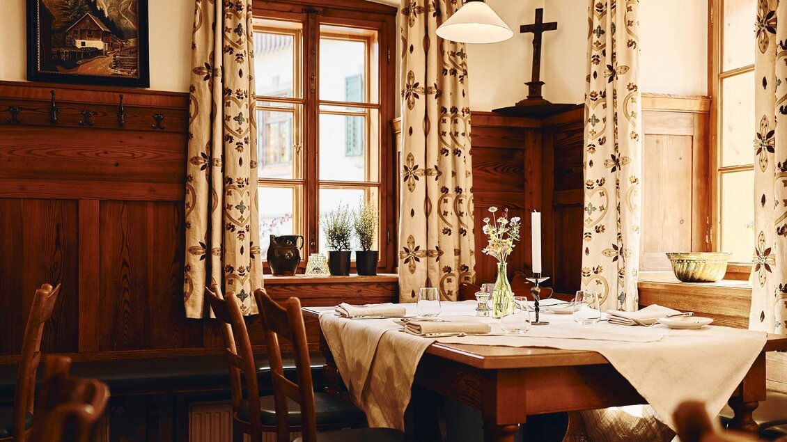 Ein gemütlicher Essbereich mit Holzmöbeln und blühenden Vorhängen. Auf dem Tisch steht eine kleine Vase mit Blumen und der Raum hat große Fenster mit viel Tageslicht. | © Armin Walcher