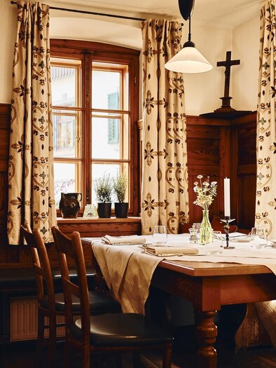 A cozy dining area with wooden furniture and blooming curtains. A small vase with flowers is on the table, and the room has large windows with plenty of daylight. | © Armin Walcher