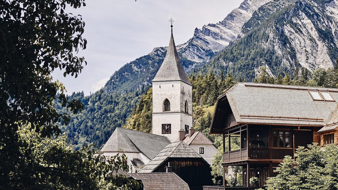 Ein malerisches Dorf mit einer Kirche und traditionellen Holzhäusern, umgeben von majestätischen Bergen. Die ländliche Kulisse strahlt Ruhe und Frieden aus. | © Armin Walcher