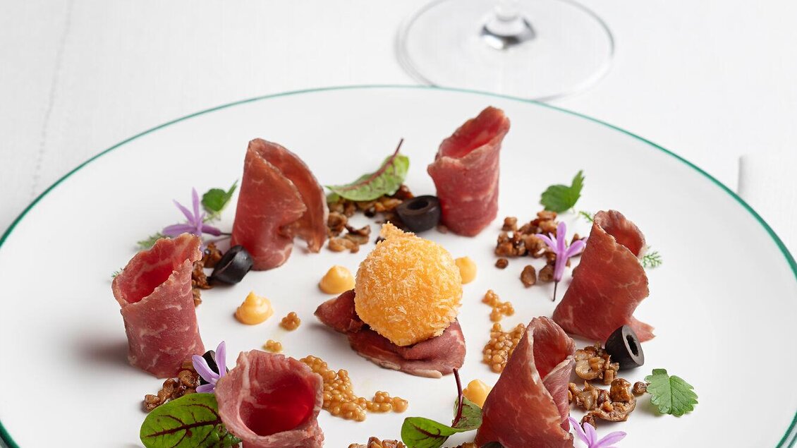 Ein eleganter Teller mit feinem Aufschnitt, umgeben von Kräutern und knusprigen Beigaben. Im Hintergrund steht ein Glas Weißwein. | © TAUROA GmbH