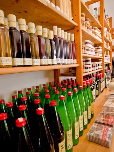 Ein gemütlicher Laden mit Regalen voller Flaschen und Gläser. Auf der linken Seite stehen grüne und rote Flaschen, während eine Person im Hintergrund interessiert schaut.