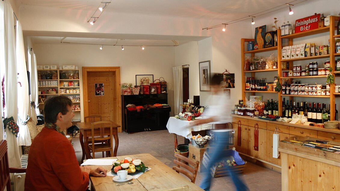 Ein gemütlicher Innenraum mit einem Holztisch und Regal voller Getränke und Lebensmittel. Eine Person bewegt sich im Hintergrund, während eine andere am Tisch sitzt und etwas schreibt.