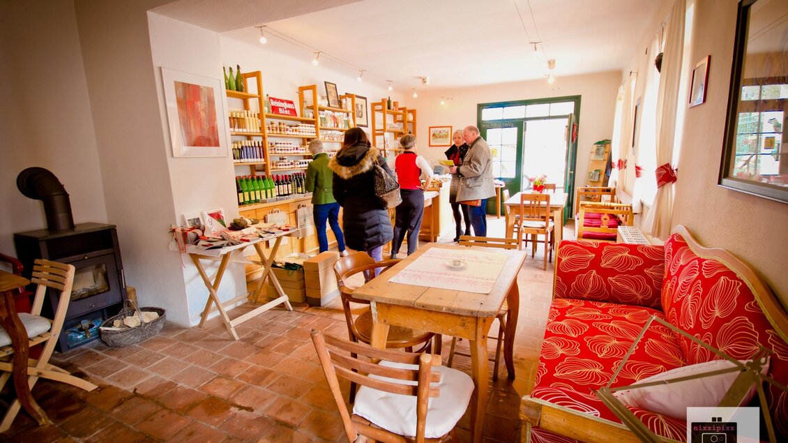 Ein gemütliches Café mit roten Sofas und Holztischen. Es gibt Regale mit Produkten und mehrere Personen, die sich im Raum unterhalten.