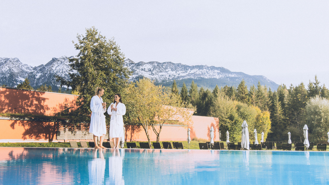Zwei Frauen in Bademänteln stehen am Rand eines blauen Pools. Im Hintergrund sind Berge und Bäume sichtbar. | © Aldiana GmbH