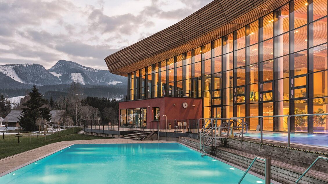 Ein modernes Gebäude mit großen Fenstern und einem einladenden Pool. Im Hintergrund sind schneebedeckte Berge und eine ruhige Landschaft zu sehen. | © GrimmingTherme / P.Aldag
