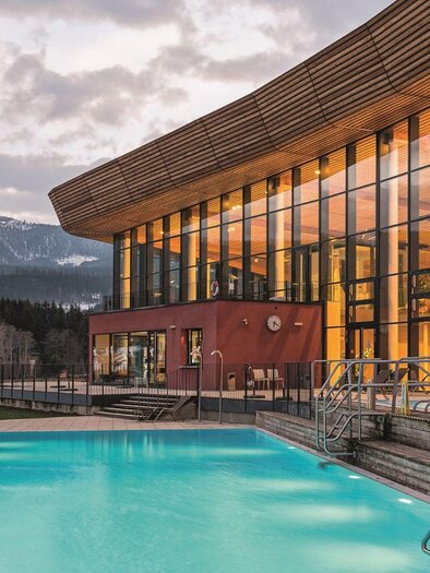 Ein modernes Gebäude mit großen Fenstern und einem einladenden Pool. Im Hintergrund sind schneebedeckte Berge und eine ruhige Landschaft zu sehen. | © GrimmingTherme / P.Aldag