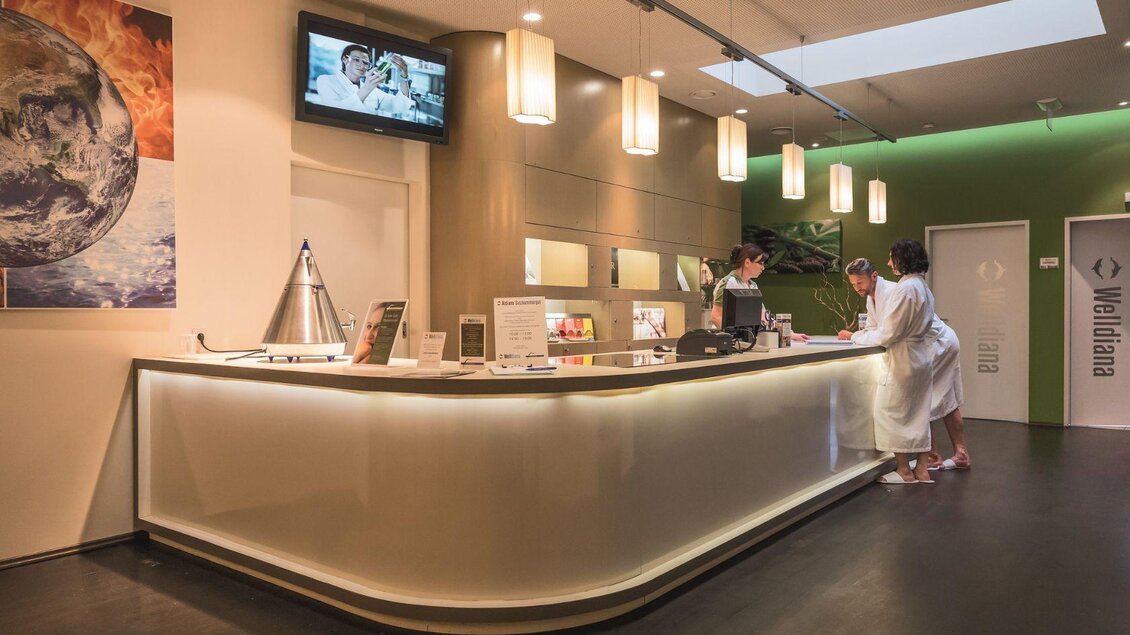 Ein eleganter Empfangsbereich mit einem modernen Tresen und sanfter Beleuchtung. Zwei Personen, eine im Bademantel, sprechen an der Rezeption. | © GrimmingTherme / P.Aldag