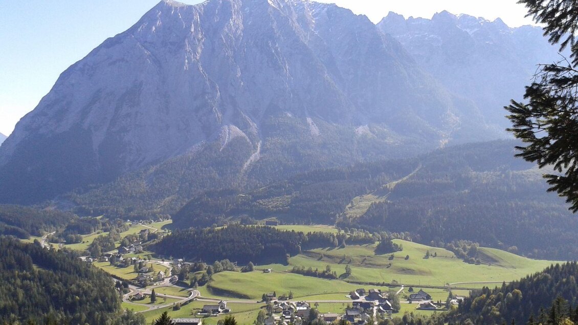 Eine eindrucksvolle Berglandschaft mit hohen Gipfeln und weitläufigen Wiesen. Im Vordergrund ist ein malerisches Dorf in einem grünen Tal zu sehen.