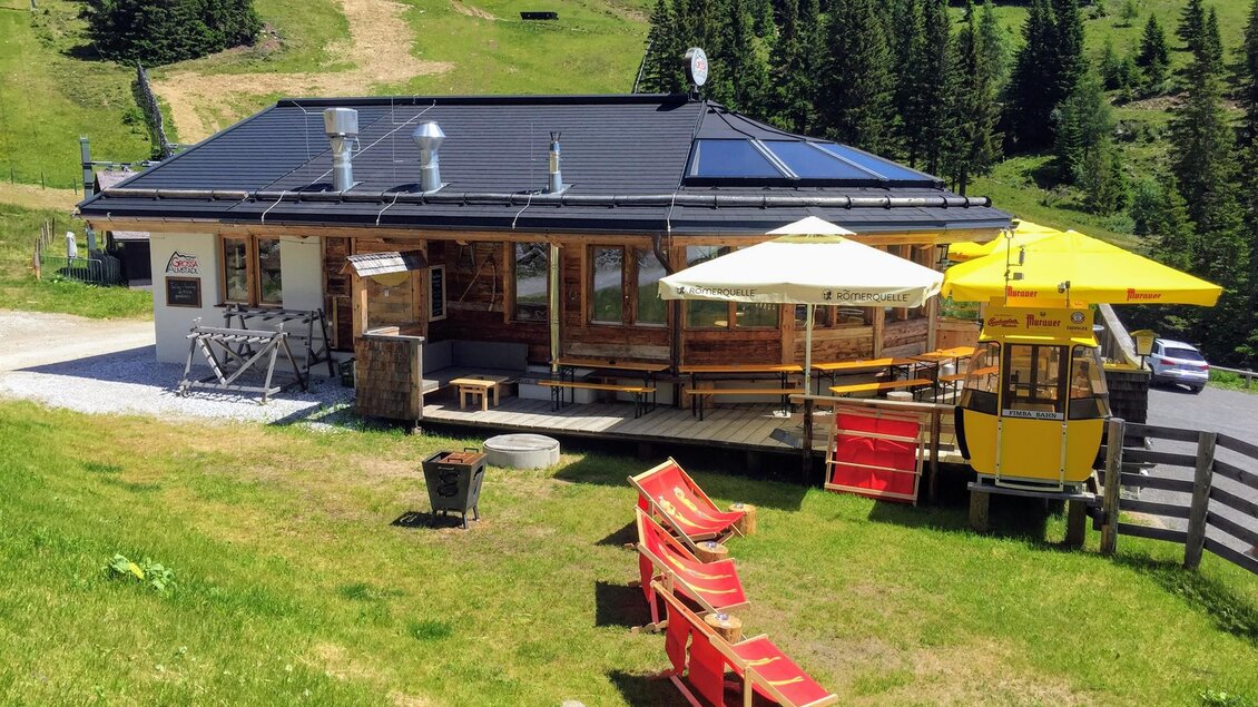 Eine gemütliche Berghütte mit einer Terrasse und Sonnenschirmen. Im Hintergrund sind grüne Wiesen und bewaldete Hügel zu sehen.