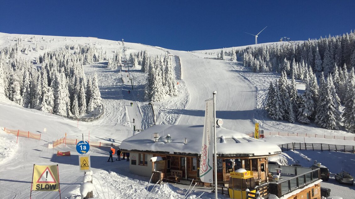 Ein schneebedeckter Skiort mit einem charmanten Holzgebäude. Im Hintergrund sind verschneite Berge und blauer Himmel zu sehen.