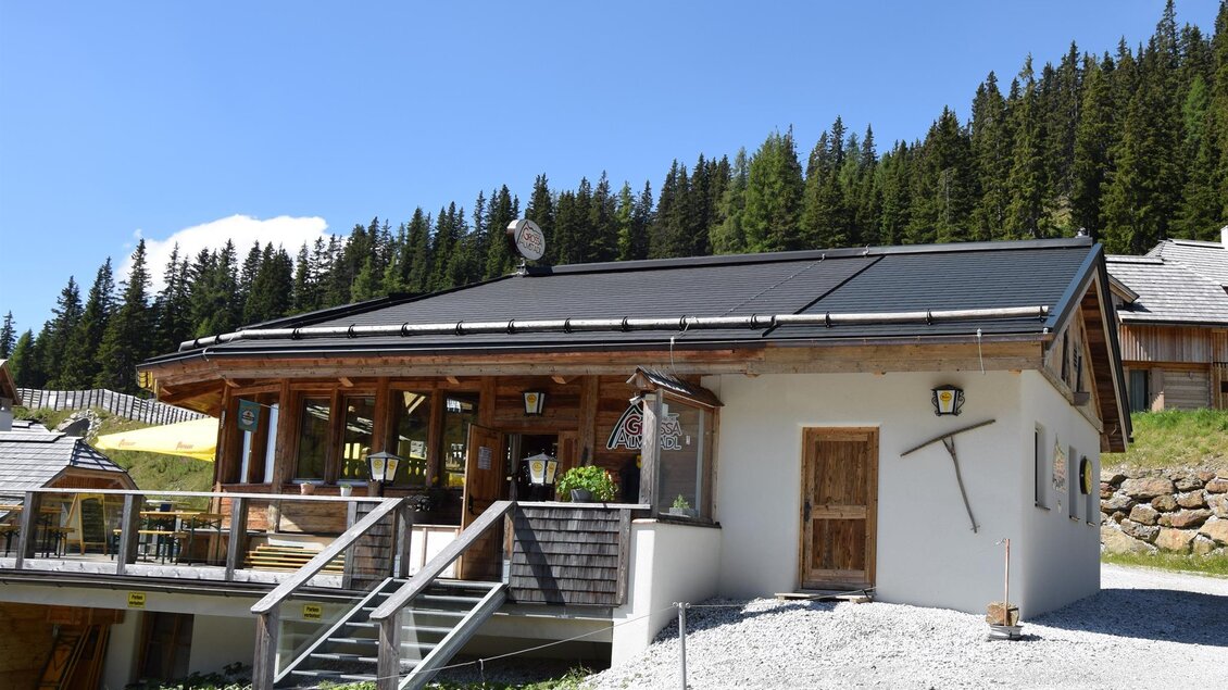 Ein charmantes Gebäude im alpinen Stil mit Holzverkleidung und einem schattigen Vordach. Umgeben von grünen Bäumen und Bergen, ideal für eine gemütliche Auszeit.