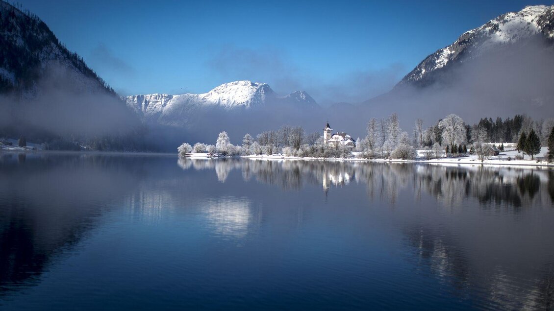 Eine malerische Winterlandschaft mit einem ruhigen See und schneebedeckten Bergen im Hintergrund. Der Himmel ist klar und blau, und der Schnee reflektiert sanft das Licht.