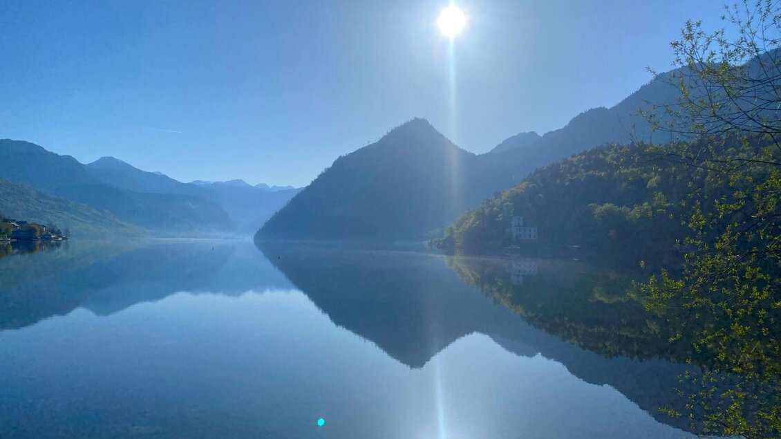 Ein ruhiger See umgeben von Bergen unter einem blauen Himmel. Die Sonne spiegelt sich im Wasser und erzeugt eine friedliche Atmosphäre. | © Bettina Scheck