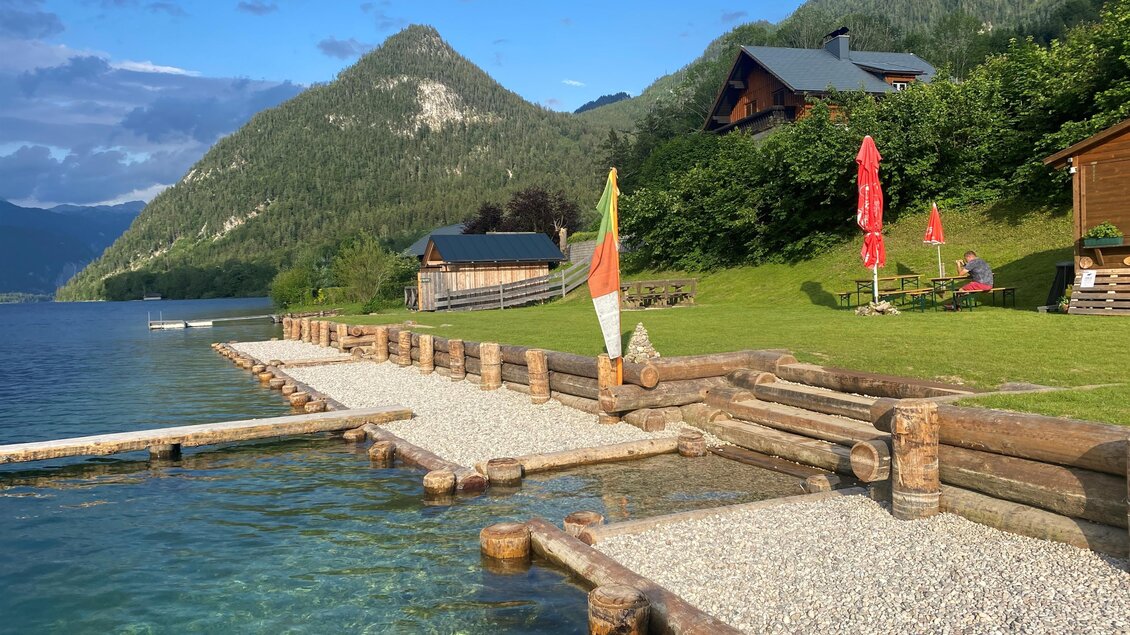 Ein schöner See mit klaren Wasser und einem Holzsteg. Im Hintergrund sind grüne Berge und einige Ferienhäuser zu sehen. | © Bettina Scheck
