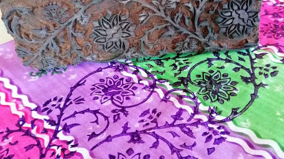Ein handgeschnitzter Block aus Holz liegt auf einem bunten Stoff mit floralen Mustern in Pink, Lila und Grün. Die Kombination der Farben und das detaillierte Design verleihen dem Bild eine kreative Atmosphäre. | © Nora Schönfellinger