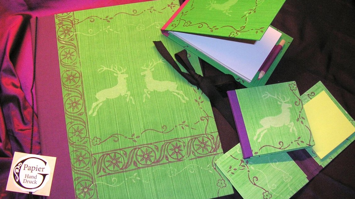 Ein Set aus buntem Papier mit grünem Hintergrund und weißen Hirschmotiven. Es besteht aus einem großen Notizbuch, mehreren Blöcken und einem Binder. | © Nora Schönfellinger