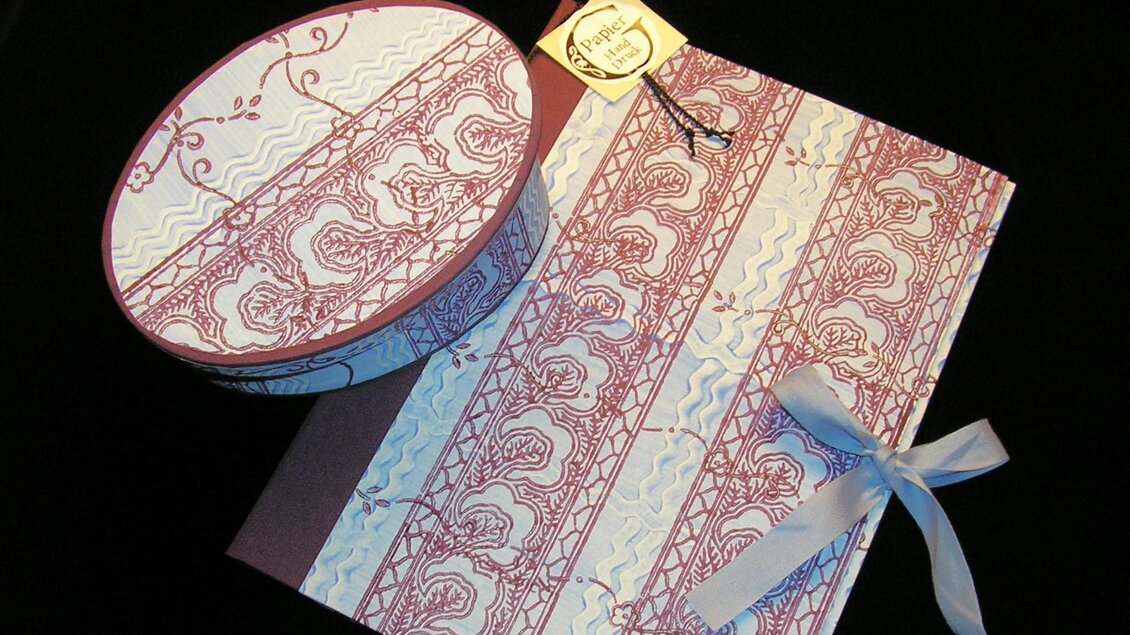 Eine dekorative Geschenkbox und ein Notizbuch mit einem eleganten Muster in Rot und Weiß. Die Box ist oval und das Notizbuch hat eine schöne Schleife. | © Nora Schönfellinger