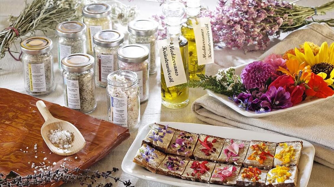 Eine ansprechende Anordnung von Gewürzen in Gläsern, Ölflaschen und frischen Blumen. Im Vordergrund sind kunstvoll dekorierte Quadrate mit essbaren Blüten zu sehen.