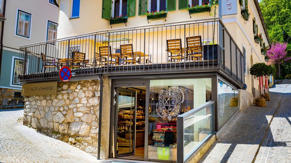 Ein charmantes Geschäft mit einem modernen Eingang und einem Außenbereich mit Holztischen. Der Weg führt sanft an dem Gebäude mit grünen Fensterläden vorbei. | © Gut Schlossberg