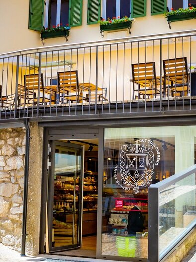 A charming shop with a modern entrance and an outdoor area with wooden tables. The pathway gently leads past the building with green window shutters. | © Gut Schlossberg