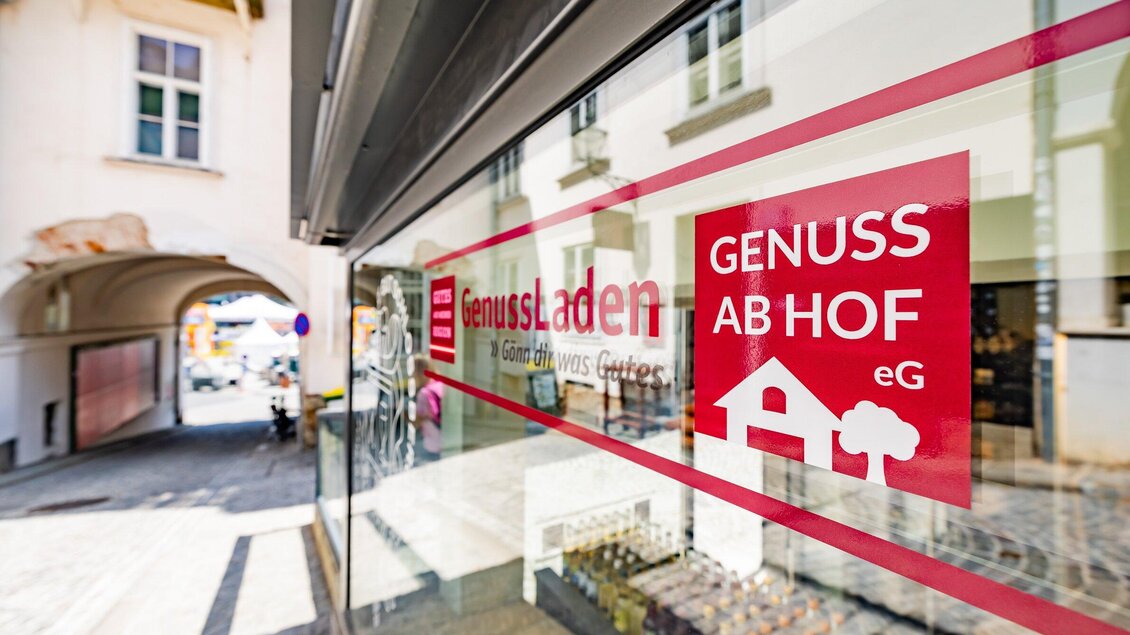 Ein Geschäft mit dem Namen "GenussLaden" ist im Bild zu sehen. Das Schaufenster zeigt ein einladendes Design und einen Zugang zu einer charmanten Straße. | © Gut Schlossberg