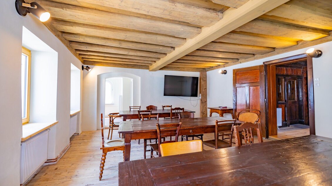 Ein heller Raum mit Holzvertäfelung und mehreren Tischen und Stühlen. An einer Wand befindet sich ein Fernseher und große Fenster sorgen für viel Tageslicht. | © Gut Schlossberg