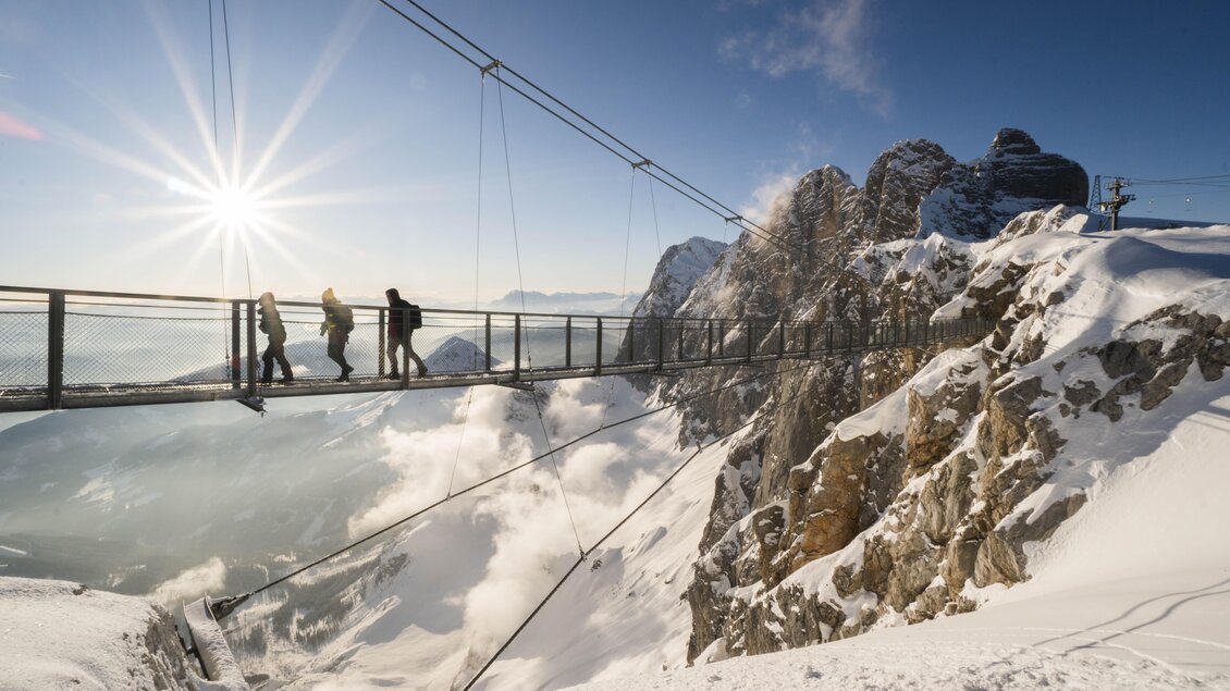 Eine schneebedeckte Berglandschaft mit einer schwebenden Hängebrücke. Menschen gehen auf der Brücke und genießen die Aussicht auf die Wolken und die Berge. | © David McConaghy