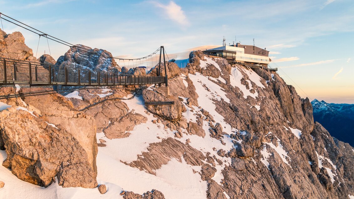 Eine beeindruckende Berglandschaft mit schneebedeckten Felsen und einem Gebäude auf dem Gipfel. Eine Hängeseilbrücke verbindet die Felsen und bietet eine atemberaubende Aussicht. | © Alexander Klünsner