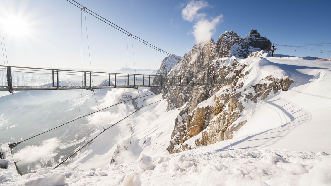 Eine schneebedeckte Berglandschaft mit einer Seilbrücke, die über die Klippen führt. Die Sonne scheint klar am Himmel und es gibt Wolken in der Ferne. | © David McConaghy