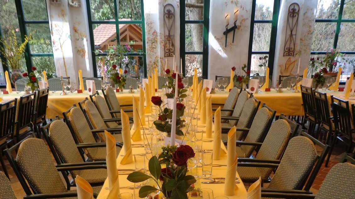 Lange Tafel mit gelber Decke, Stoffservietten und Blumenschmuck vor Fenstern. | © Häuserl im Wald - Legenstein
