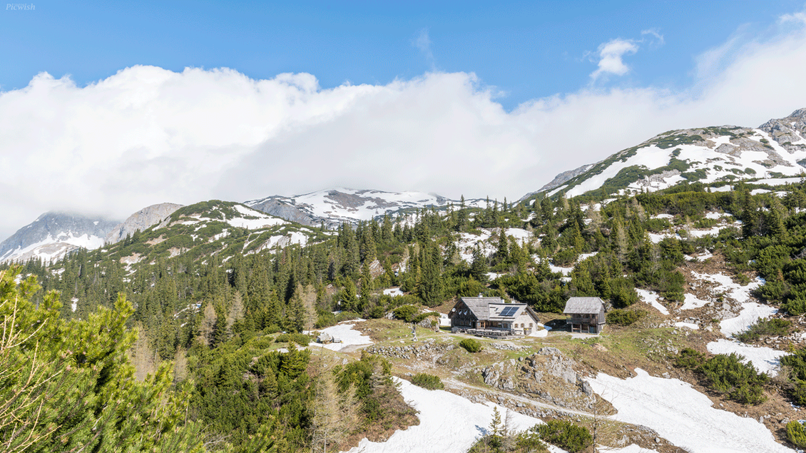 Eine malerische Berglandschaft mit schneebedeckten Gipfeln und grünen Wäldern. Zwischen den Bergen steht eine charmante Hütte.