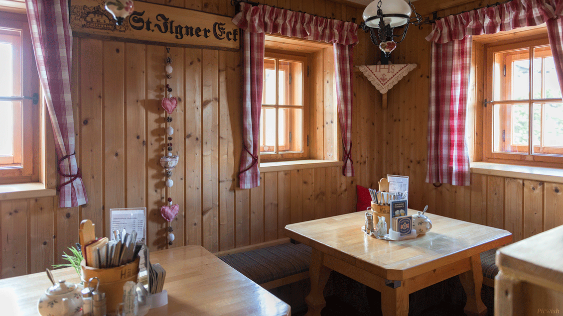 Eine gemütliche Stube mit Holzwänden und traditionellen Fenstern. Der Tisch ist liebevoll eingedeckt und es gibt dekorative Elemente wie Herzchen und Pflanzen.