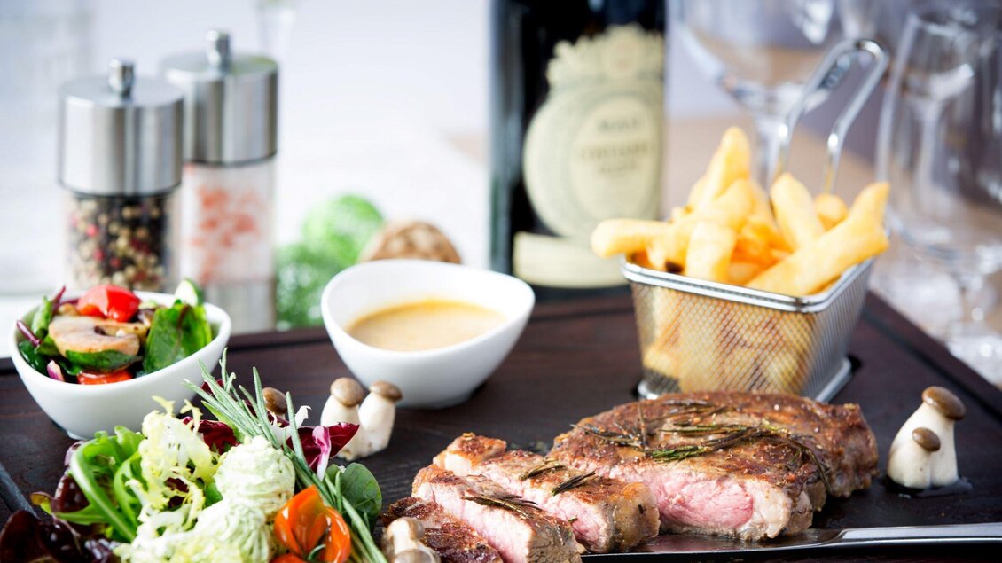 Ein köstliches Steakgericht mit Beilagen wie Pommes frites und einem frischen Salat. Im Hintergrund steht eine Flasche Wein und Gewürzmühlen. | © Huegellandhof
