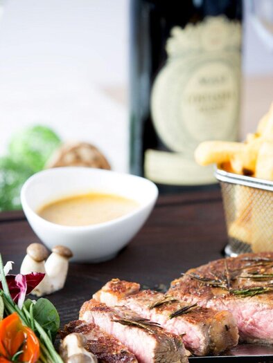 Ein köstliches Steakgericht mit Beilagen wie Pommes frites und einem frischen Salat. Im Hintergrund steht eine Flasche Wein und Gewürzmühlen. | © Huegellandhof
