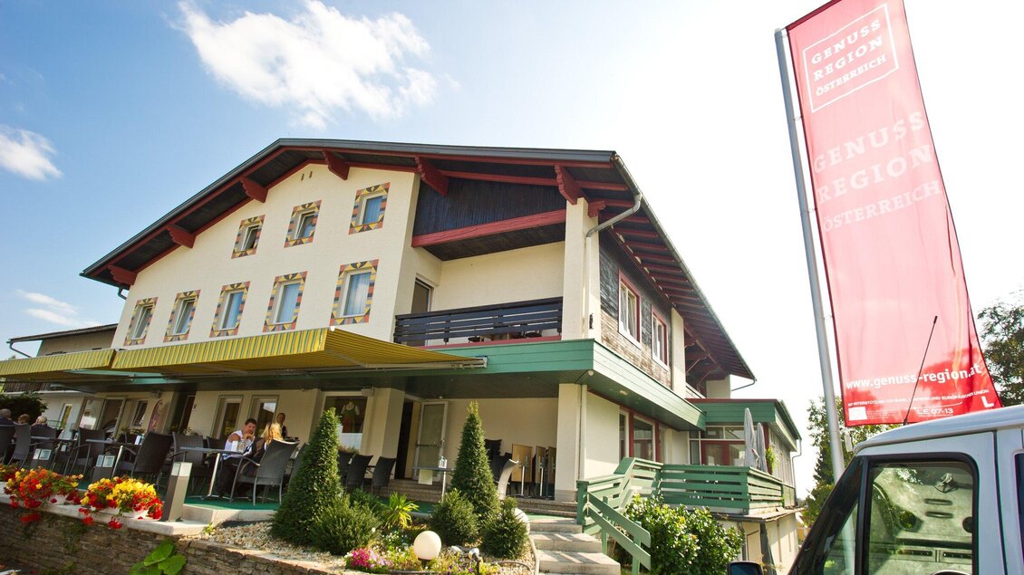 Ein modernes Gebäude mit einem Restaurantbereich und Terrasse. Vor dem Haus sind blühende Blumen und ein Werbeschild sichtbar. | © Huegellandhof
