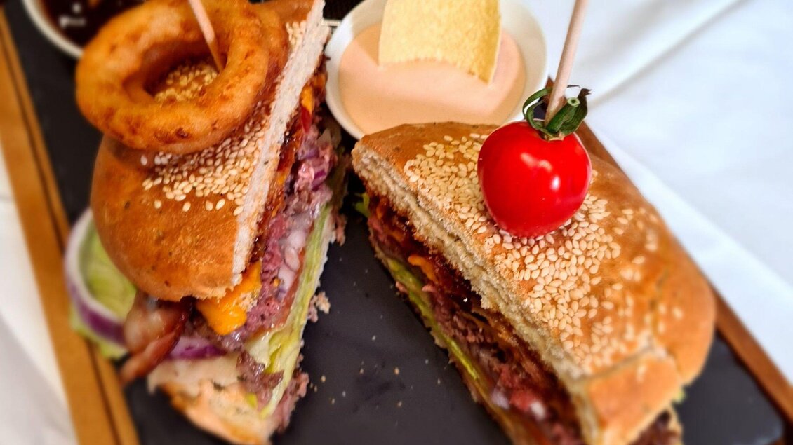 Ein köstlicher Burger mit Sesambrötchen, gefüllt mit saftigem Fleisch, frischem Gemüse und Käse. Daneben liegt eine Portion Zwiebelringe und eine kleine Sauce. | © Huegellandhof