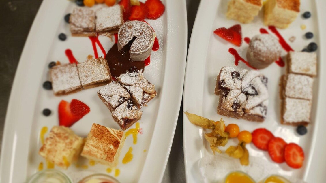 Zwei Platten mit verschiedenen Desserts sind zu sehen. Die Süßspeisen sind mit Puderzucker bestäubt und mit frischen Früchten und Saucen dekoriert. | © Huegellandhof