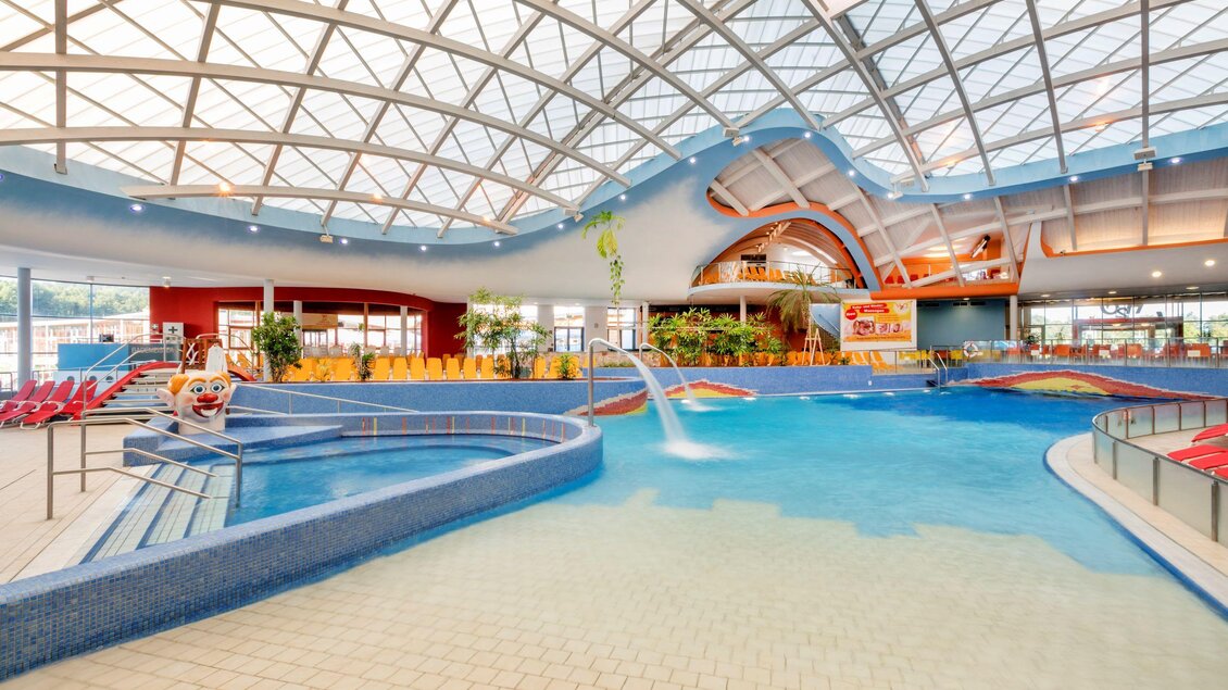 Ein modernes Hallenbad mit gläsernem Dach und blauen Wasser. Es gibt Rutschen und Liegen für Besucher. | © H2O Hotel-Therme-Resort