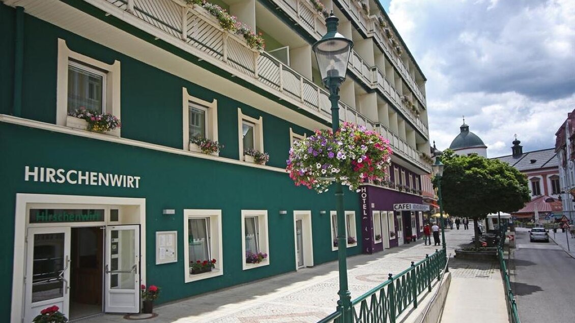 Eine bunte Straße mit einem grünen Gebäude und Blumenkästen. Im Hintergrund sind Wolken und ein Kirchturm sichtbar. | © www.weisser-hirsch.at