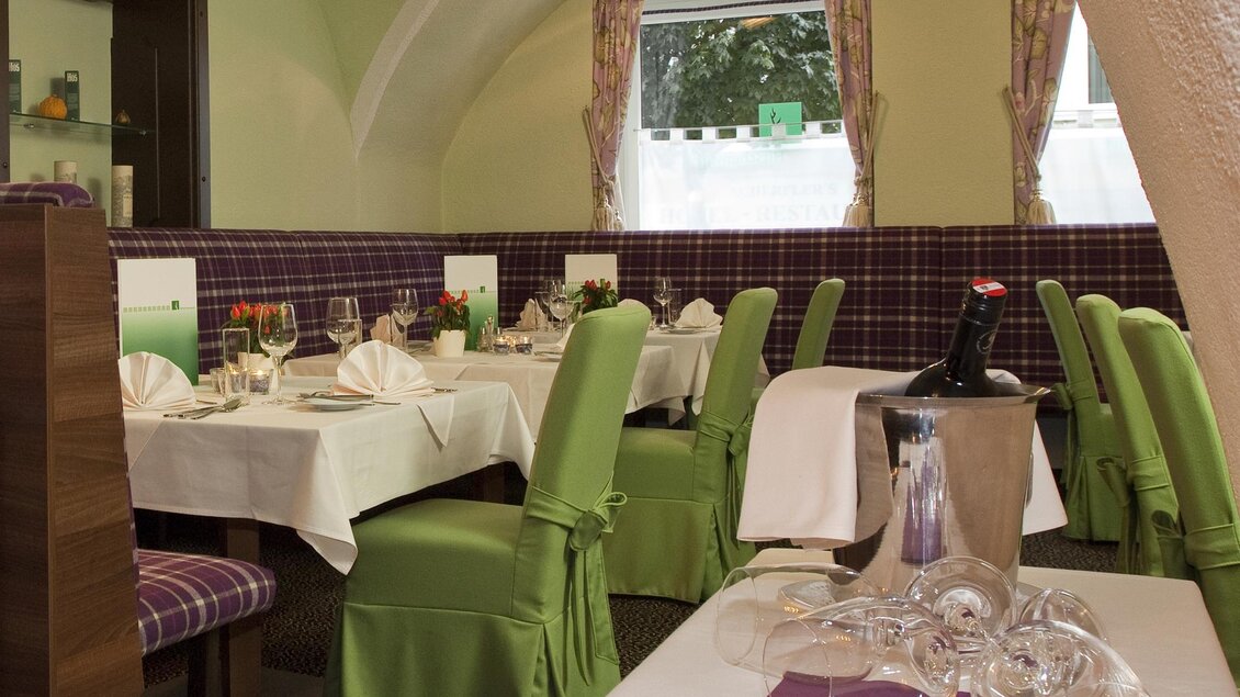 Ein eleganter Restaurantbereich mit Tischen, die mit weißen Tischdecken und grünen Stühlen gedeckt sind. Sanfte Beleuchtung und große Fenster schaffen eine einladende Atmosphäre. | © www.weisser-hirsch.at