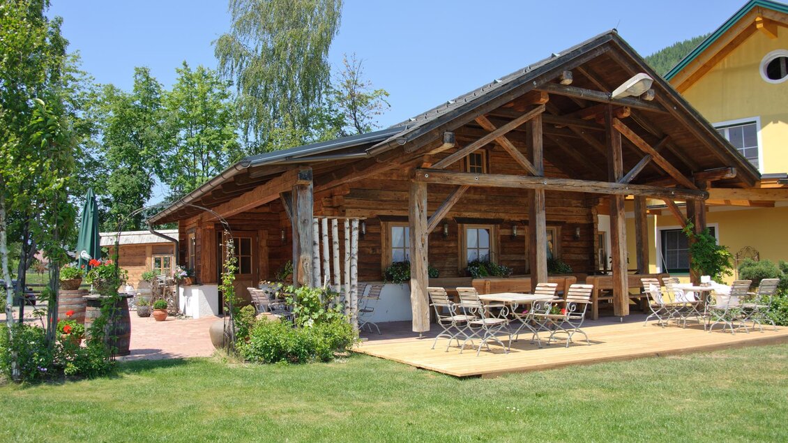 Ein gemütliches Holzhaus mit einer überdachten Veranda und Sitzgelegenheiten im Freien. Umgeben von grünen Bäumen und einem sonnigen Himmel. | © Haigls Hofschenke