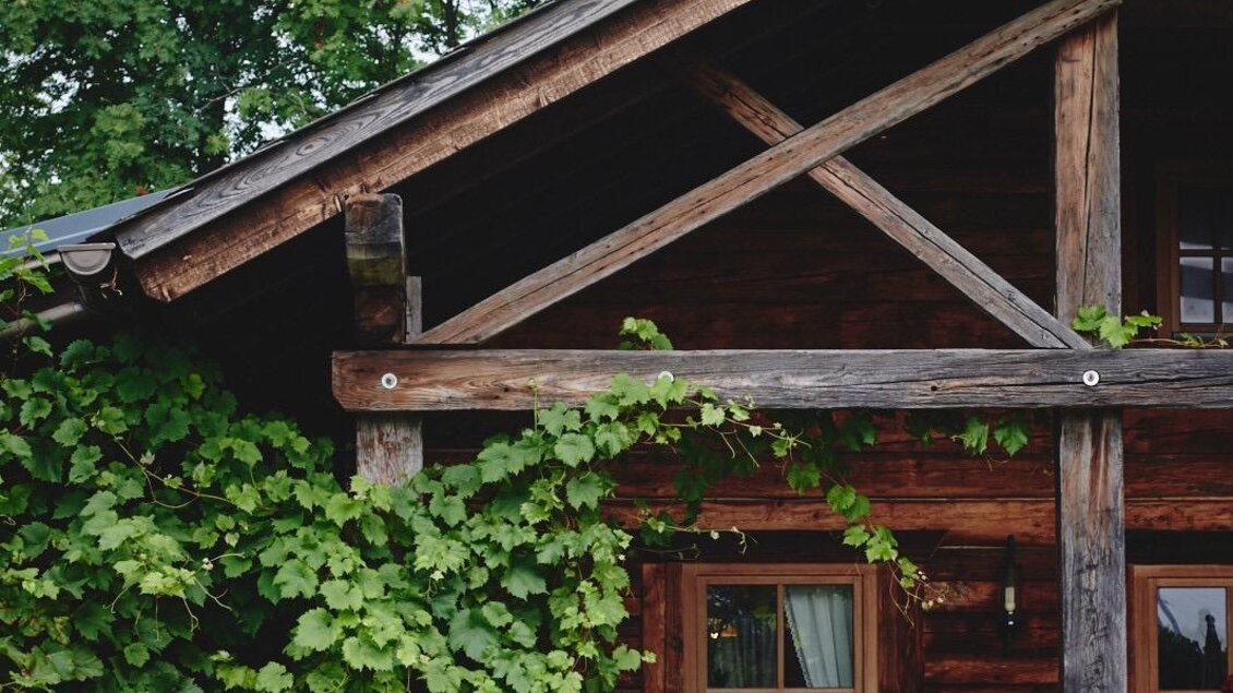 Ein rustikales Holzhaus mit üppigem Grün und blühenden Pflanzen. Ein gedeckter Tisch mit Stühlen lädt zum Verweilen ein. | © Haigls Hofschenke