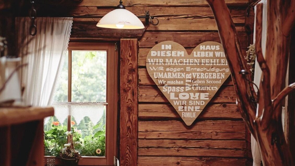 Ein gemütlicher Raum aus Holz mit einem Tisch und Stühlen. An der Wand hängt ein herzförmiges Dekor mit einem liebevollen Spruch. | © Haigls Hofschenke