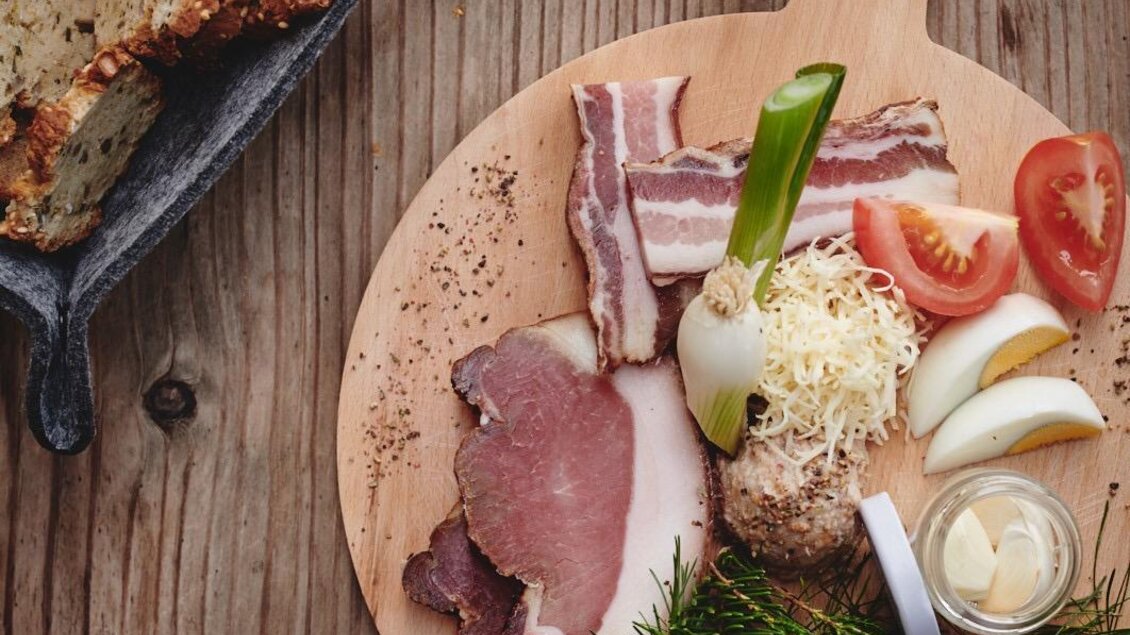 Ein rustikales Holzbrett mit einer Auswahl an Wurst und Fleisch, garniert mit Gemüsestücken und Kräutern. Daneben befindet sich ein Korb mit Brot. | © Haigls Hofschenke